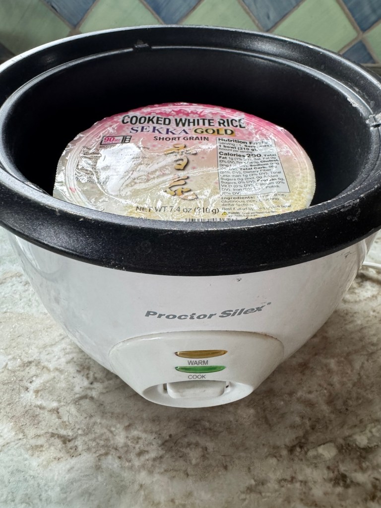 A Proctor Silex rice cooker containing a sealed package of cooked white rice labeled 'Sekka Gold Short Grain'.